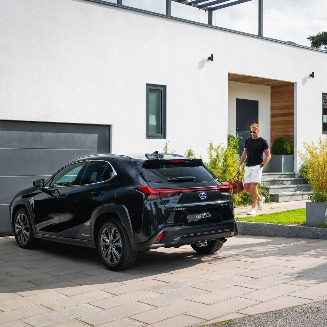 Blonde haired man walks from front door to a parked UX 250h in the driveway. He is carrying a bag and wearing sunglasses.