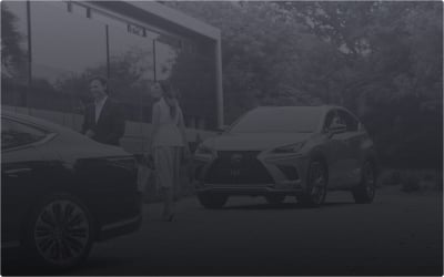 Two people stand between a black sedan and a silver Lexus NX SUV parked outdoors.